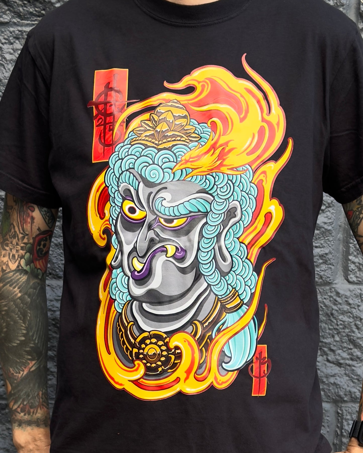 Black t-shirt with a colorful graphic design of a mask and flames on a stone wall background.